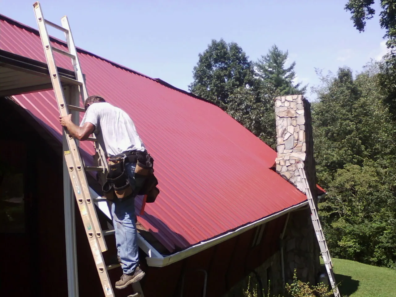 Roofer on a ladder beside a red metal roof and stone chimney — Soffit & Fascia Repair by Roofing Brothers in Winneconne